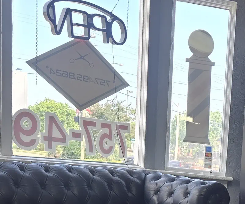 The front room: a black tufted Chesterfield sofa under the bay window, with the shop's phone number, the OPEN sign, the hanging diamond sign and a barber pole in the windows